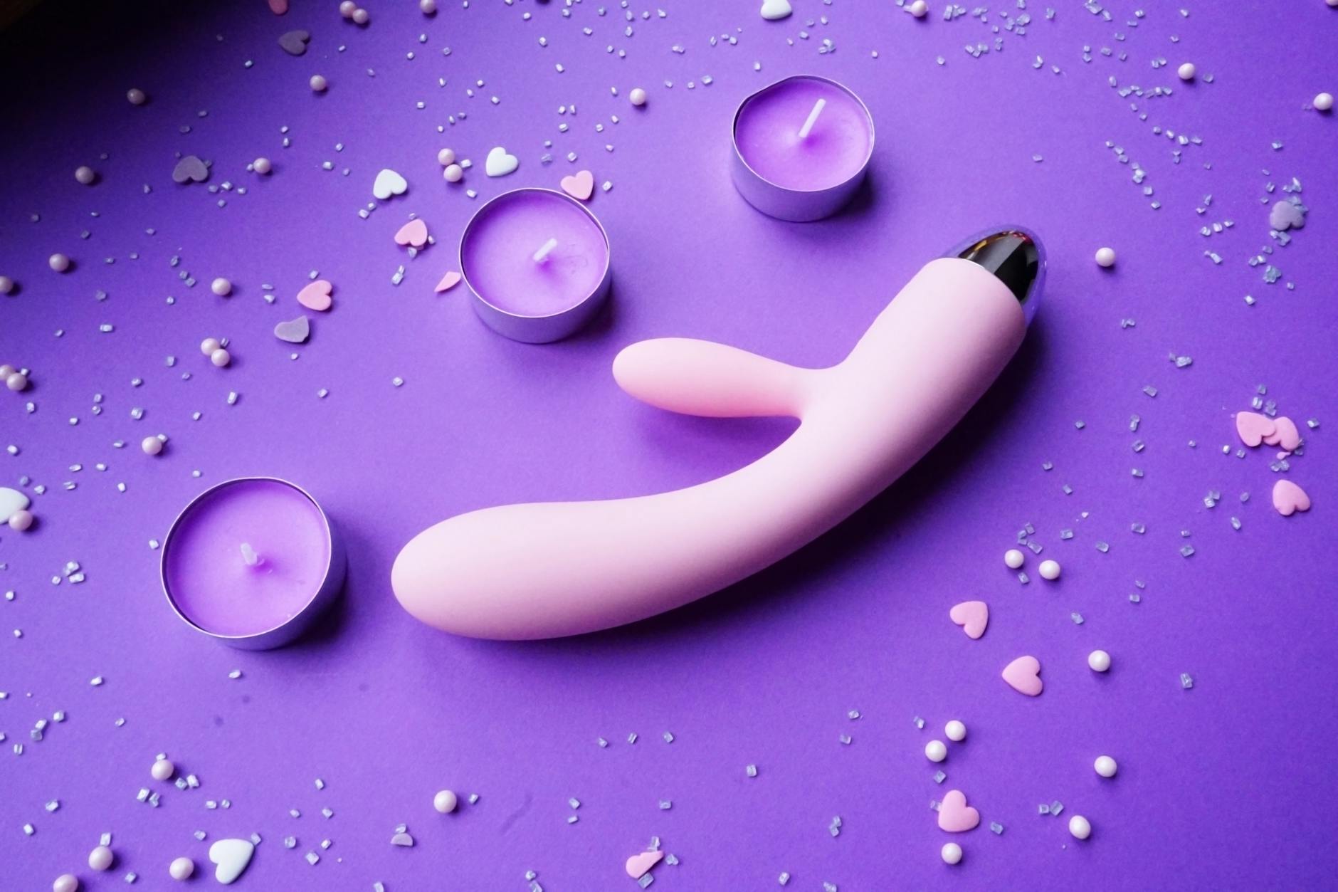 Pink vibrator surrounded by candles and heart confetti on a purple background, symbolizing intimacy and self-love.